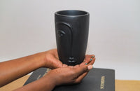 Person holding a black ceramic mug with a face design on a plain background