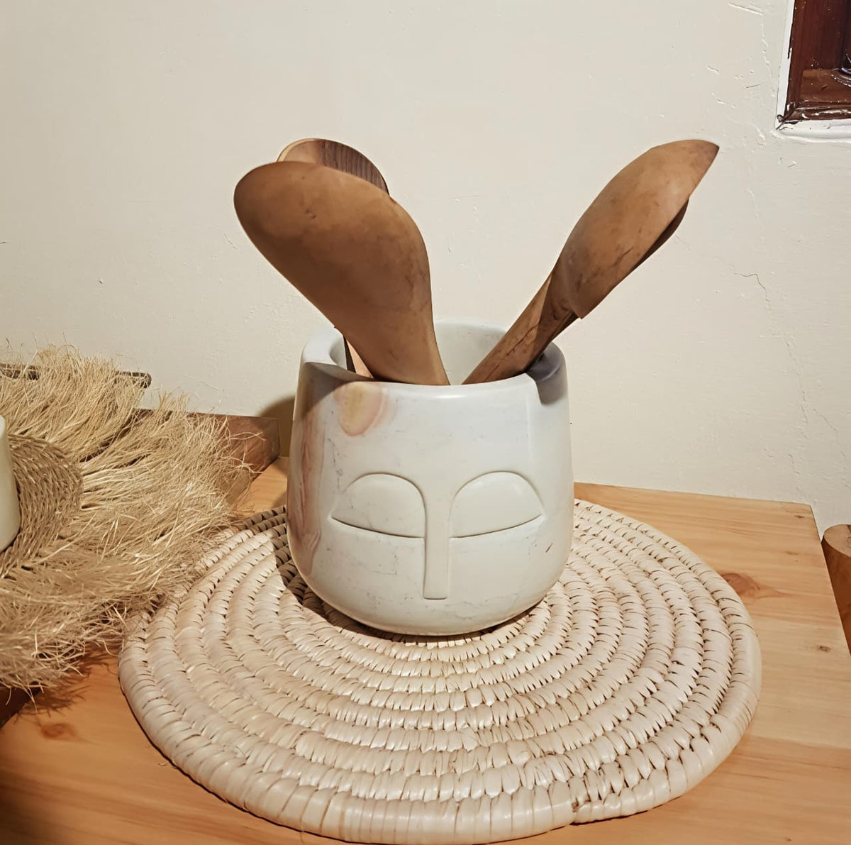 Ceramic container with face design holding wooden spoons on a woven mat.