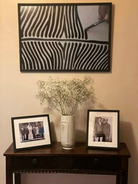 Two framed photographs on a wooden table with a vase containing small white flowers, against a beige wall with a zebra print picture frame.