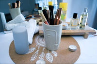 White ceramic cup with makeup brushes on a decorative mat with candles and a bottle in the background.