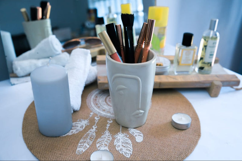 White ceramic cup with makeup brushes on a decorative mat with candles and a bottle in the background.