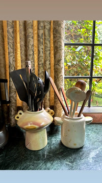 Two ceramic utensil holders with kitchen utensils on a table with a window and curtain in the background.