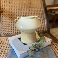 White ceramic vase with two handles on a stack of books with green leaves, against a woven chair background.