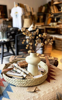 Decorative setting with a vase of dried flowers on a woven tray in a room with furniture and decor.