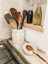 Wooden utensils in a white container on a kitchen counter with bottles in the background.