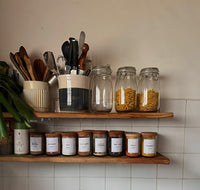 Kitchen shelves with jars, utensils, and spice containers against a tiled wall.