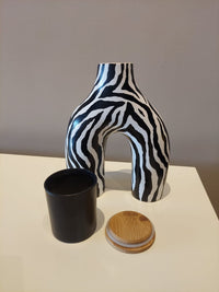 Zebra-striped vase with a black cup and wooden lid on a light surface.