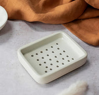 Rectangular ceramic soap dish with holes on a light gray surface with an orange cloth in the background.